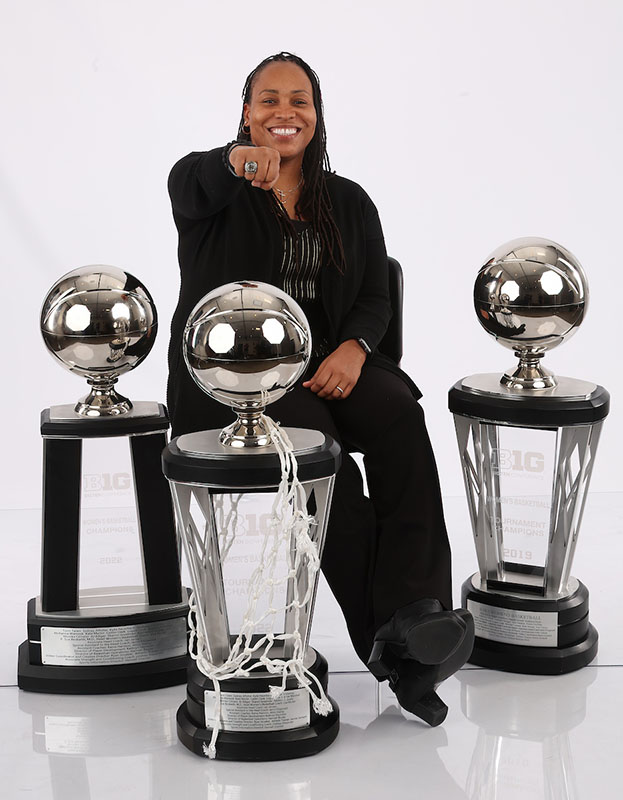 Coach Harmon sits surrounded by trophies and shows her championship ring to the camera
