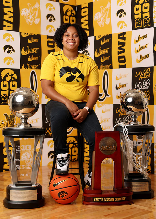 A woman is seated, surrounded by basketball trophies