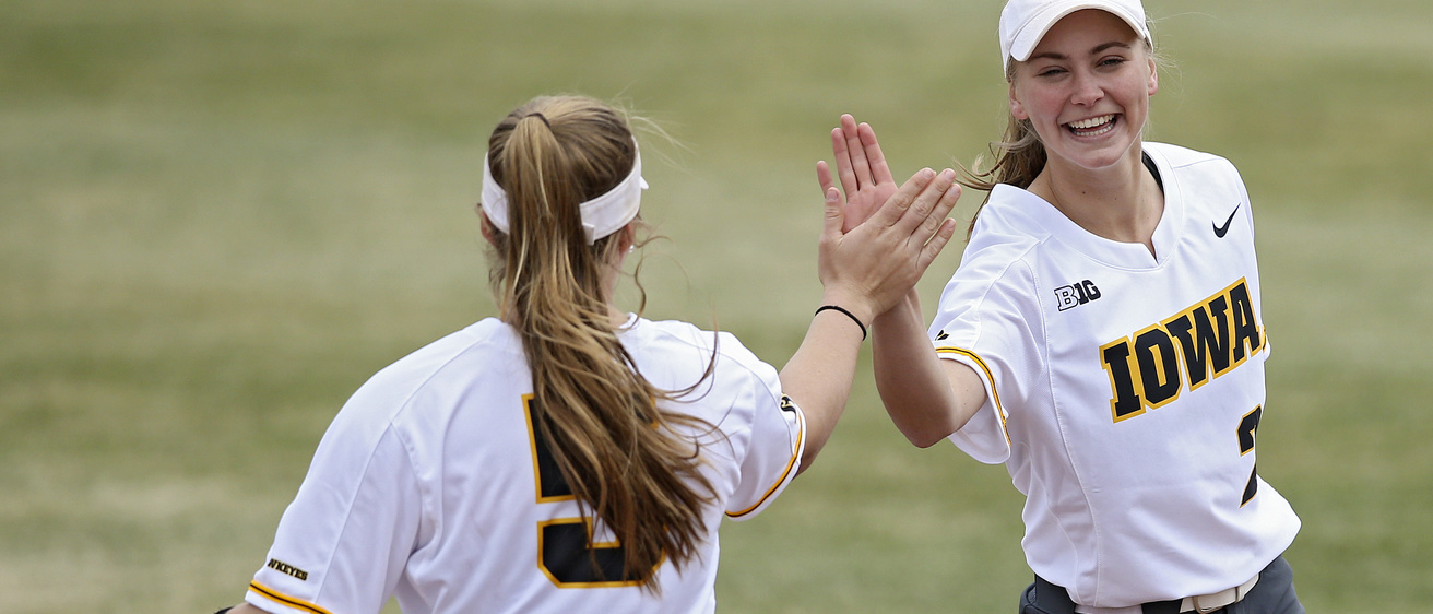 Two softball players wearing white uniforms exchange a high five